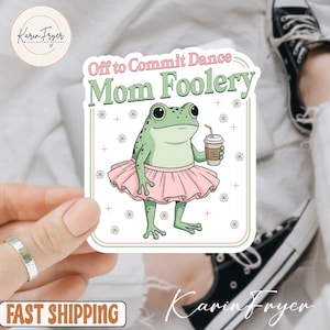 May include: A sticker featuring a cartoon frog wearing a pink tutu and holding a coffee cup. The text "Off to Commit Dance Mom Foolery" is above the frog. The sticker has a white background with a green and pink border.