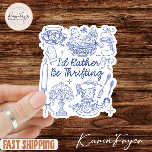 May include: A white sticker with blue line art illustrations of kitchen and tea party items, including a chicken, teacups, and a rolling pin. The text "I'd Rather Be Thrifting" is in the center. The sticker is held by a hand.