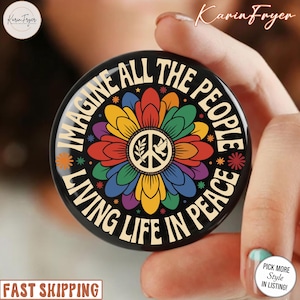 May include: A black round phone grip with the words "IMAGINE ALL THE PEOPLE LIVING LIFE IN PEACE" in white. The center features a colourful flower with a peace symbol and a dove. The grip is held by a person.