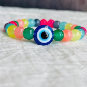 May include: A colorful beaded bracelet with a blue evil eye charm. The beads are in shades of pink, yellow, green, and blue.