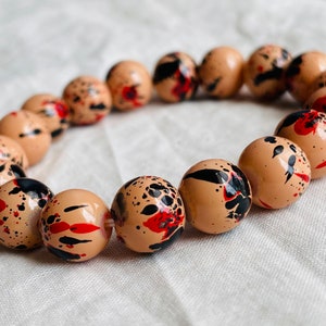 May include: A beige bracelet with black and red splatter paint design. The bracelet is made of round beads.