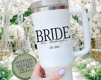May include: White insulated tumbler with a handle, featuring the word "BRIDE" in black, along with "Mrs. Washington - EST. 2026." The tumbler has a silver rim and a clear lid. The background shows a floral wedding arch.