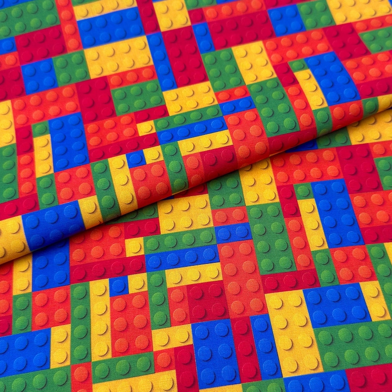 May include: A colorful fabric with a repeating pattern of toy building blocks in red, yellow, green, and blue. The blocks are arranged in a grid-like design, creating a playful and vibrant aesthetic. The fabric is likely suitable for various craft projects.