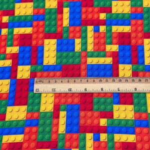 May include: A colorful fabric featuring a pattern of interlocking toy blocks in red, yellow, green, and blue. The blocks are arranged in a grid-like pattern, with a ruler showing measurements in centimeters.