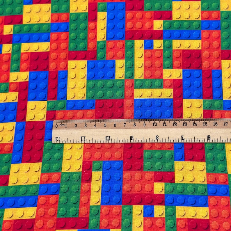 May include: A colorful fabric featuring a pattern of interlocking toy blocks in red, yellow, green, and blue. The blocks are arranged in a grid-like pattern, with a ruler showing measurements in centimeters.