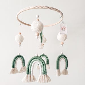 May include: A handmade baby mobile featuring a wooden hoop with a macrame design. The mobile includes white yarn pom-poms, wooden beads, and green and white yarn rainbows. A small tag with a rainbow graphic is attached.