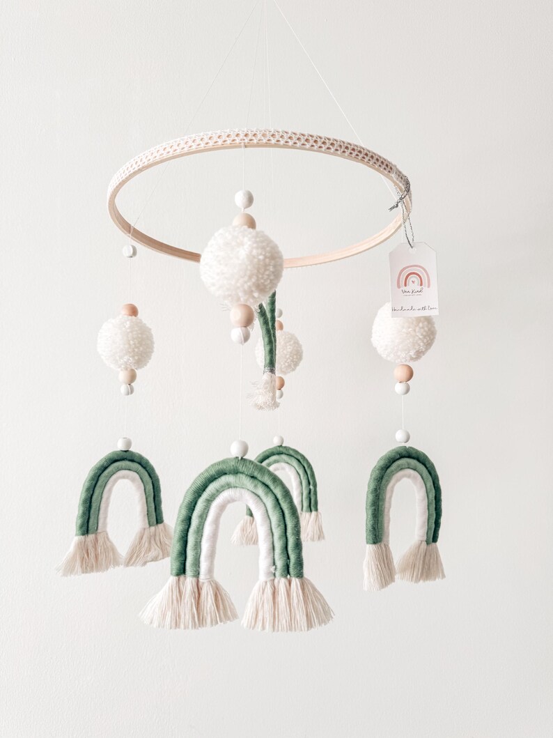 May include: A handmade baby mobile featuring a wooden hoop with a macrame design. The mobile includes white yarn pom-poms, wooden beads, and green and white yarn rainbows. A small tag with a rainbow graphic is attached.