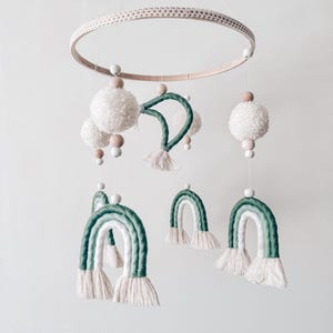May include: A handmade baby mobile featuring a wooden ring with a woven texture. The mobile includes white pom-poms, wooden beads, and green and white rainbow-shaped decorations, all suspended by strings.