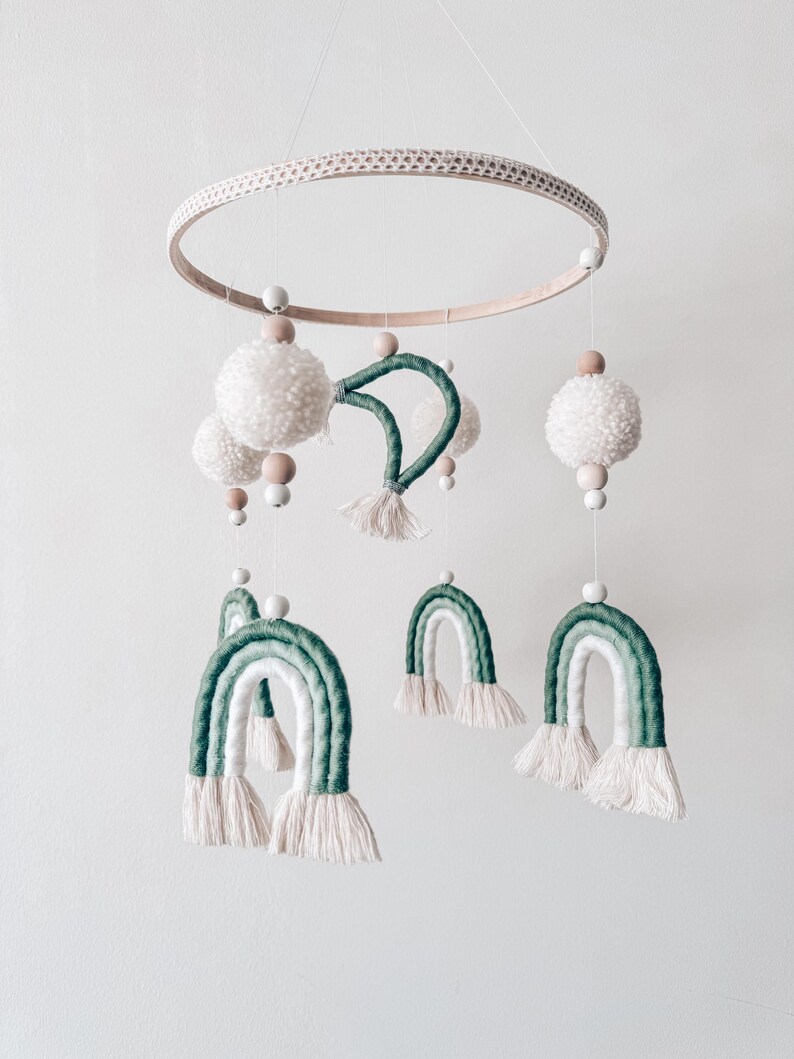 May include: A handmade baby mobile featuring a wooden ring with a woven texture. The mobile includes white pom-poms, wooden beads, and green and white rainbow-shaped decorations, all suspended by strings.
