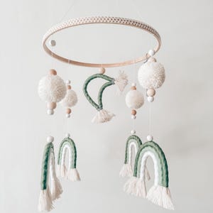 May include: A handmade baby mobile featuring a wooden ring with a woven texture, adorned with white pom-poms, wooden beads, and green and white rainbow-shaped accents. The mobile is suspended by a thin white string, creating a whimsical nursery decoration.