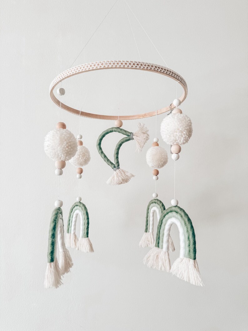 May include: A handmade baby mobile featuring a wooden ring with a woven texture, adorned with white pom-poms, wooden beads, and green and white rainbow-shaped accents. The mobile is suspended by a thin white string, creating a whimsical nursery decoration.