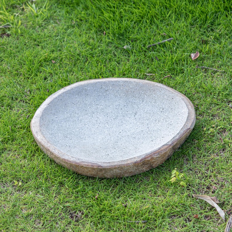 May include: A round, gray stone bird bath with a rough, textured surface. The bird bath is sitting on a bed of green grass.