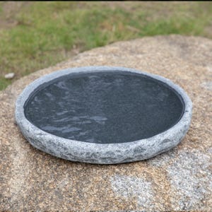 Op de afbeelding: Een ronde, grijze stenen vogelbad met een ruw, getextureerd oppervlak. Het bad is gevuld met water.