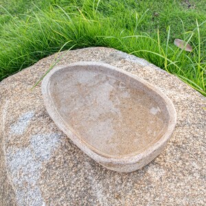 May include: A light brown oval-shaped stone bird bath sits on a larger, rough-textured gray stone. The bird bath is empty and appears to be made of natural stone.