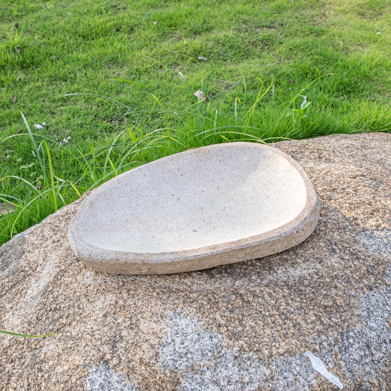 May include: A white oval-shaped stone bird bath with a rough texture. The bird bath is sitting on a large, brown, textured rock.