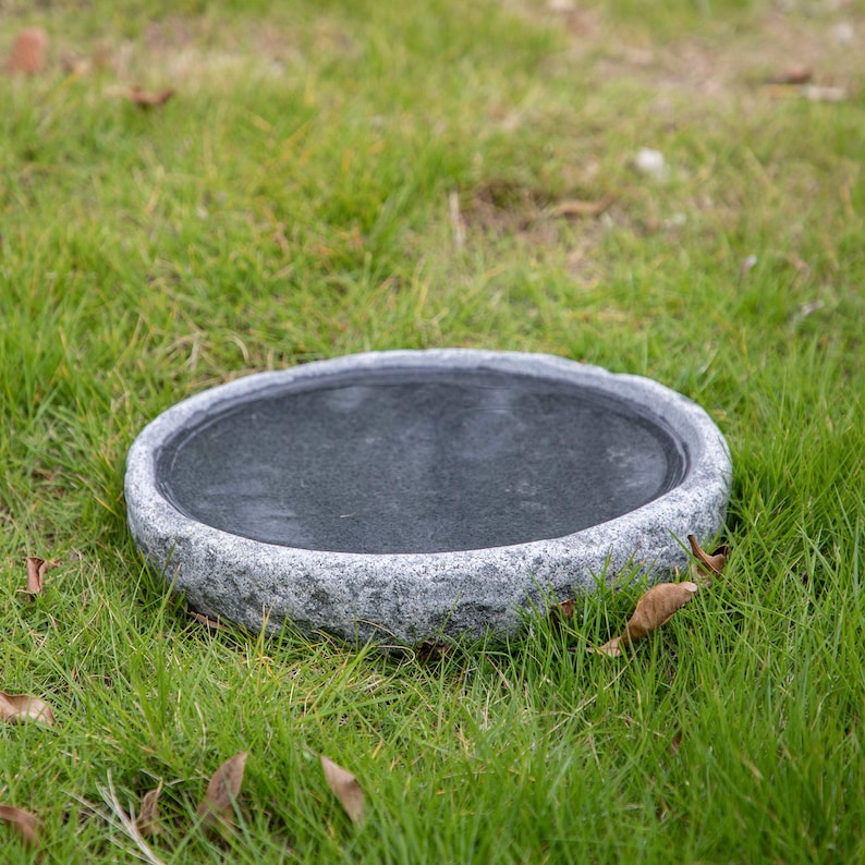 Op de afbeelding: Een ronde, grijze stenen vogelbad met een glad, zwart interieur. Het vogelbad staat op een bed van groen gras.