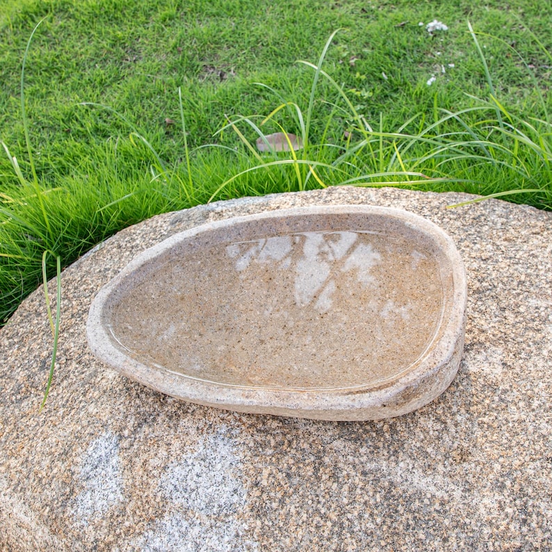 May include: A light brown stone bird bath with a shallow basin, sitting on a larger, rough-textured stone. The basin is filled with water, reflecting the surrounding greenery.