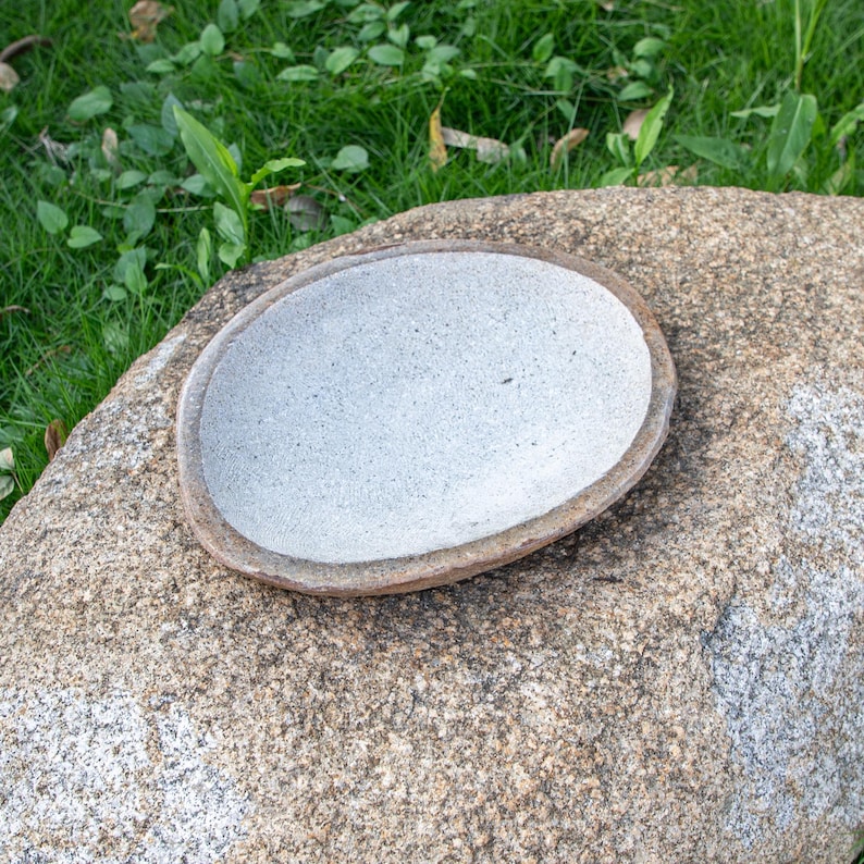 May include: A white ceramic bowl with a brown rim sits on a large, gray rock. The bowl is slightly oval-shaped and has a rough, textured surface.