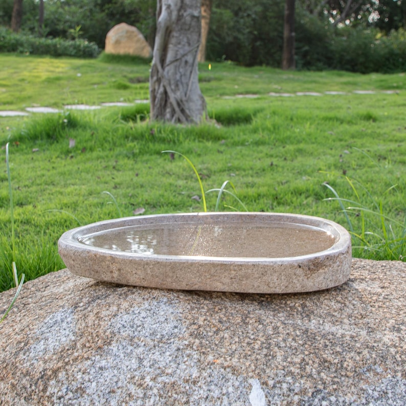 May include: A light brown oval-shaped stone bird bath with water in it, sitting on a large gray rock in a grassy garden.