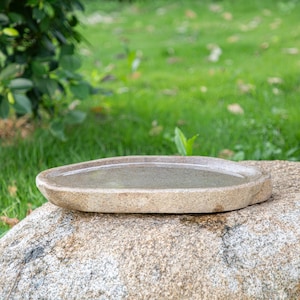 May include: A light brown oval-shaped stone bird bath sits on a large gray rock. The bird bath is filled with water.