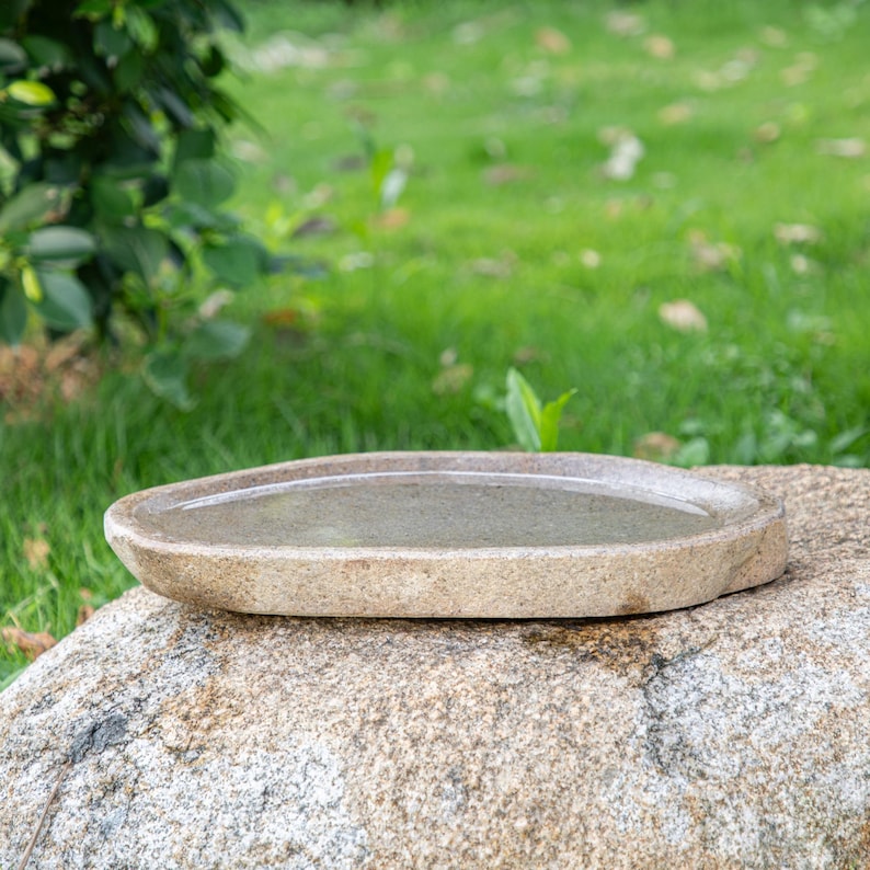 May include: A light brown oval-shaped stone bird bath sits on a large gray rock. The bird bath is filled with water.