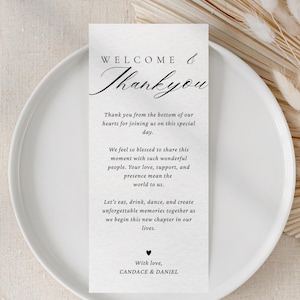 以下が含まれることがあります： 「ようこそ＆ありがとう」と書かれたエレガントな筆記体の白いウェディングメニューカードが白いお皿の上に置かれています。カードには心温まるメッセージが書かれており、ドライフラワーのアクセントが添えられています。