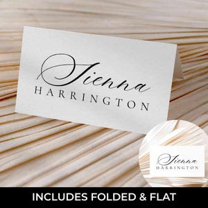 Puede incluir: Una tarjeta de mesa blanca con el nombre "Sienna Harrington" en elegante escritura negra. La tarjeta está doblada y descansa sobre una superficie clara y texturizada. El texto "INCLUDES FOLDED & FLAT" está en la parte inferior.