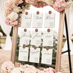 May include: A gold framed seating chart with nine cards, each labeled with a table number in elegant calligraphy. The cards are affixed with wax seals. The frame is decorated with pink and white flowers, creating a romantic aesthetic.