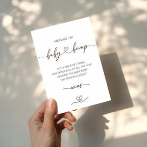 Può includere: Una carta bianca con testo nero che recita "MEASURE THE baby & bump". La carta include le istruzioni per un gioco di baby shower. Le parole "wins" e un cuore sono in fondo alla carta.