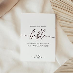 May include: A white card with the text "PLEASE SIGN BABY'S bible" in a script font. Below, it reads "HIGHLIGHT YOUR FAVORITE VERSE AND LEAVE A NOTE!" A small heart design is at the bottom. The card is on a white fabric surface.