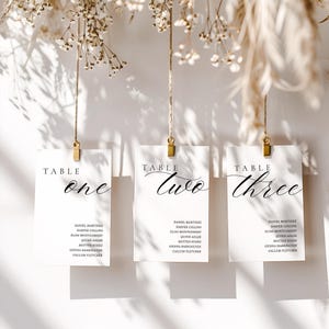 May include: Three white table cards with gold clips, each labeled "Table One," "Table Two," and "Table Three" in elegant script. Each card lists names below the table number. The cards are suspended from twine, with dried flowers in the background.