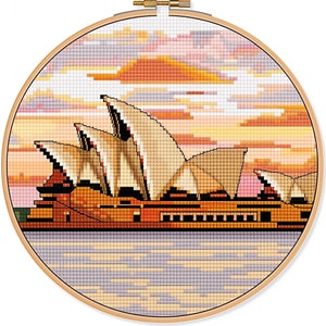 Könnte beinhalten: Eine Kreuzstich-Stickerei des Opernhauses von Sydney bei Sonnenuntergang. Das berühmte Gebäude ist in Braun-, Beige- und Weißtönen dargestellt, mit einem leuchtend orangefarbenen und rosafarbenen Sonnenuntergang im Hintergrund. Das Wasser ist in Blau- und Grautönen dargestellt.