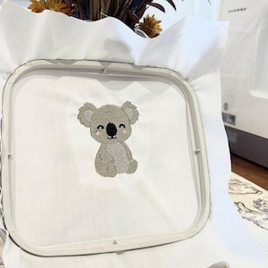 Op de afbeelding: Borduurwerk van een grijze koala met zwarte ogen en een roze neus op een witte stof in een borduurring.