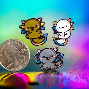 May include: Three colourful axolotl charms with rainbow tails. The axolotls are drawn in a cartoon style with big eyes and smiling mouths. The charms are made of resin and are about the size of a 20 pence coin.