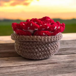 May include: A crocheted basket filled with red and white crocheted mushrooms. The basket is brown and has a textured surface. The mushrooms are arranged in a pile and have white dots on them.
