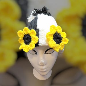 Puede incluir: Un gorro de ganchillo con un diseño a cuadros en blanco y negro, adornado con dos aplicaciones de girasoles amarillos y negros. El gorro tiene un pompón blanco y negro en la parte superior. Se exhibe en una cabeza de maniquí blanca.