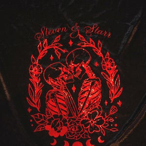 Puede incluir: Manta de terciopelo negro con un diseño de dos esqueletos abrazándose en purpurina roja, rodeados de flores y enredaderas. El texto "Steven & Starr" y "06.24.2024" también está incluido en el diseño.