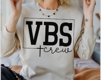 May include: A beige sweatshirt with the text "VBS crew" in black. The letters "VBS" are in a bold, rectangular font, with "crew" written in a cursive font below. A cross is between the two words.