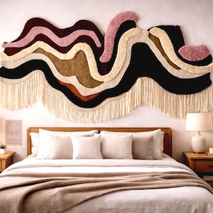 Puede incluir: Un gran tapiz de pared abstracto con un diseño en capas en tonos de negro, marrón, beige y rosa. La obra de arte textil tiene un borde con flecos y cuelga sobre una cama con ropa de cama blanca y un marco de madera. La habitación está decorada con lámparas y mesitas de noche.