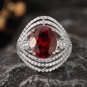 May include: A silver ring with a large oval-shaped red gemstone surrounded by a halo of smaller white gemstones. The ring has a delicate, intricate design with multiple bands of white gemstones.