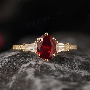 Peut inclure: Une bague en or avec une grande pierre de rubis ovale et deux diamants baguette plus petits de chaque côté. La bague est posée sur une surface texturée grise.