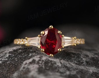 May include: A gold ring featuring a large oval ruby gemstone flanked by two smaller baguette diamonds. The ring is displayed on a grey textured surface.