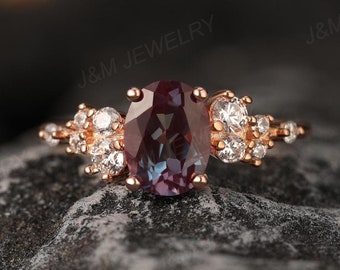 May include: A rose gold ring with a large oval-shaped alexandrite gemstone surrounded by smaller round diamonds. The ring is set on a delicate band with a cluster of smaller diamonds on each side of the center stone.