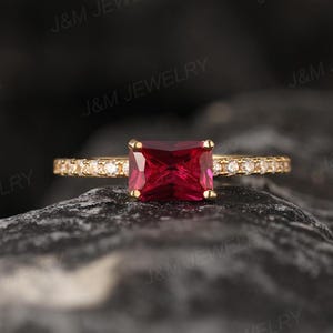 May include: A gold ring featuring a large, square, red gemstone. The band is embellished with small, clear gemstones. The ring is displayed against a dark, textured backdrop, accentuating the jewellery's sparkle and design.