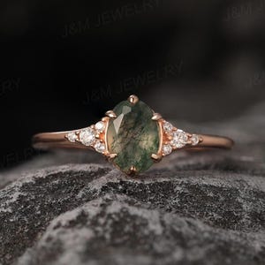 Puede incluir: Un anillo de oro rosa con una piedra de ágata musgosa en forma de marquesa. El anillo está adornado con pequeñas piedras redondas y transparentes a ambos lados de la piedra central. El anillo está sobre un fondo oscuro y texturizado.