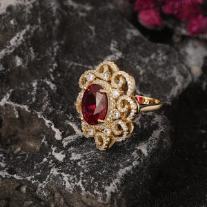 May include: A gold ring featuring a large oval ruby gemstone surrounded by smaller diamonds. The ring has a delicate, floral design.