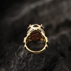 May include: A gold ring featuring a large red gemstone surrounded by smaller white gemstones. The ring has a black and gold patterned band.