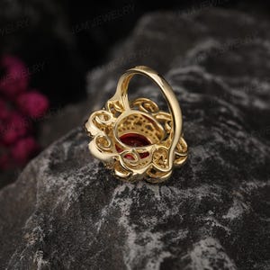 May include: A gold ring featuring a large red gemstone encircled by smaller clear gemstones. The ring boasts a delicate, intricate design.