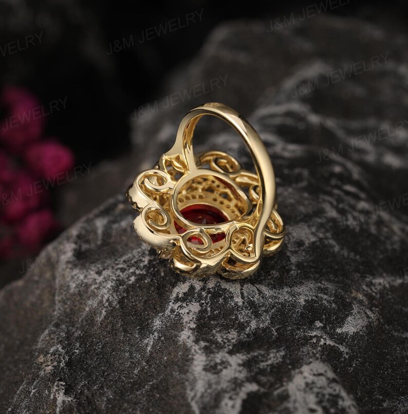 May include: A gold ring featuring a large red gemstone encircled by smaller clear gemstones. The ring boasts a delicate, intricate design.