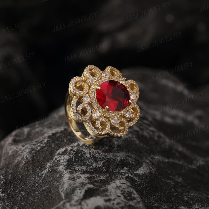 May include: A gold ring featuring a large red gemstone surrounded by smaller white gemstones. The ring has a delicate, intricate design.